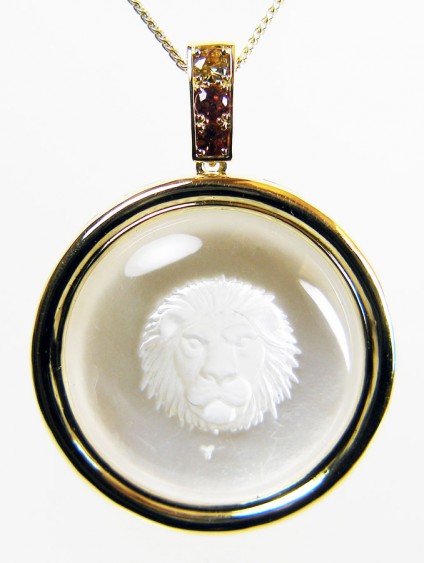 Leo pendant in rock crystal & 18ct yellow gold - Lion's head of Leo, exquisitely carved into a dome of pure rock crystal by renowned German engraver Erwin Pauly, set against a subtly shimmering background of polished mother-of-pearl and mounted in 18ct yellow gold with a line of golden Kenyan sapphires weighing 0.52ct.  Pendant is suspended from an 18" yellow 18ct gold chain. Pendant is 50mm long and 36mm wide. 